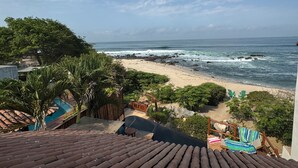 Une vue sur la plage depuis une terrasse dotée d’un toit en tuiles, surplombant une plage de sable où les vagues s’écrasent contre des rochers, le tout entouré de verdure et agrémenté de quelques espaces de détente en plein air.
