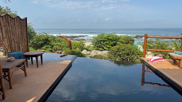 Une piscine à débordement offrant une vue sur l’océan, entourée de mobilier en bois et d’une clôture en bambou.