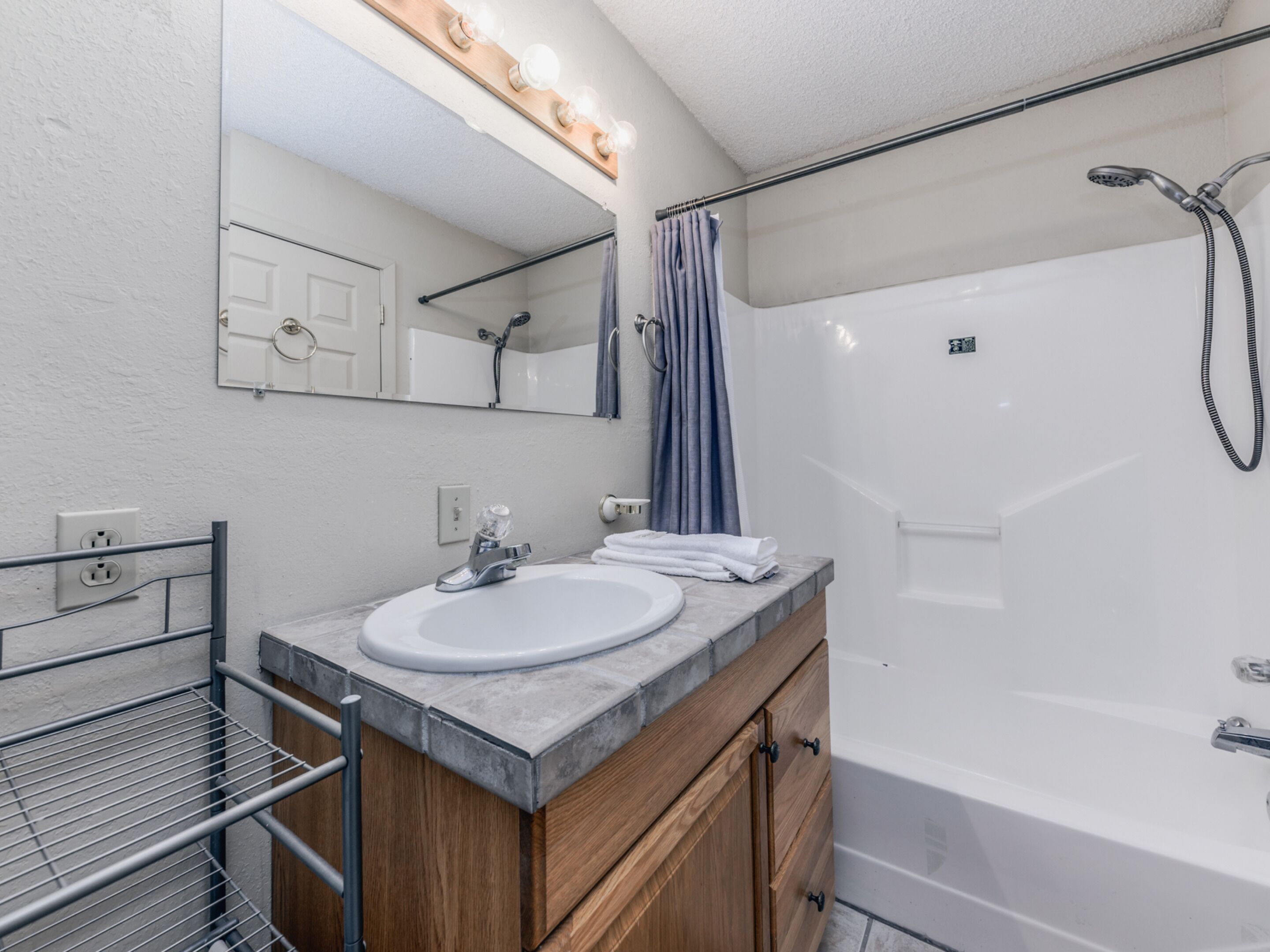 A modern bathroom with a white sink, wooden vanity, a large mirror with lights, a shower, and a towel rack.