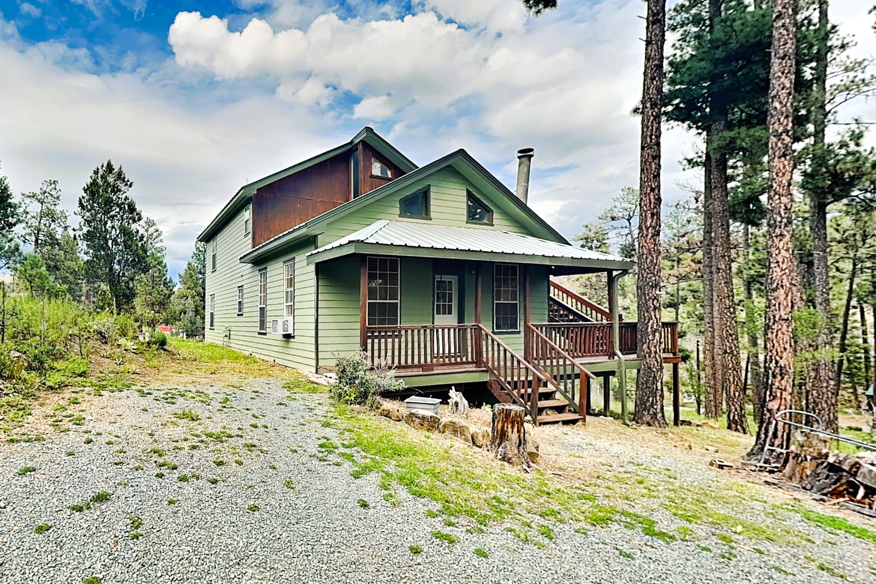 A two-story wooden house with a porch and a chimney, surrounded by trees and a gravel driveway.