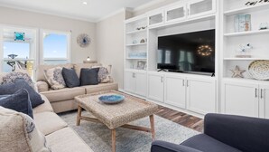 A living room with a beige sofa, a dark armchair, a woven coffee table, and a flat-screen TV on a white cabinet.
