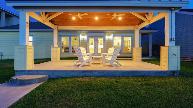 A covered patio with two Adirondack chairs and a table, a wooden ceiling, and a view of the house's interior through glass doors. - The Castaway by Miss Kitty’s (Rockport)