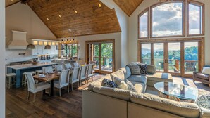 A spacious kitchen with a large island, dining area, and a cozy living room with a large window overlooking a scenic landscape. - Up & Away at Eagles Nest | Luxury Mountain Home | Sleeps 24 | Views + Hot Tub (Banner Elk)