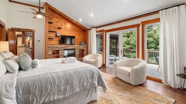 A spacious bedroom with a large bed, a fireplace, a ceiling fan, and a balcony with a view of the outdoors. - Mountaintop Ridge | Near Asheville with Pool, Fire Pit, and Ranging Views (Fairview)