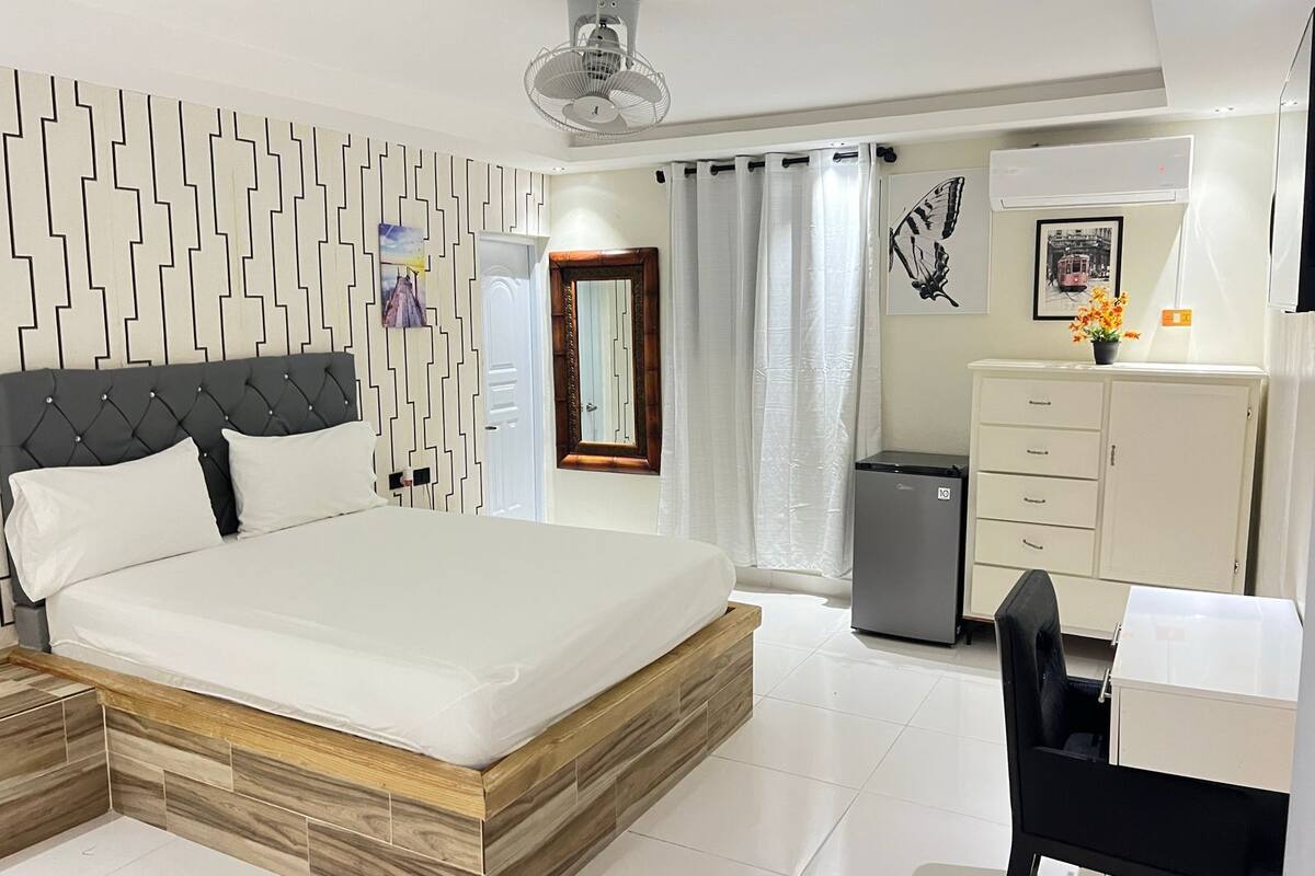 A modern hotel room with a bed, a desk, a wardrobe, and a small refrigerator.