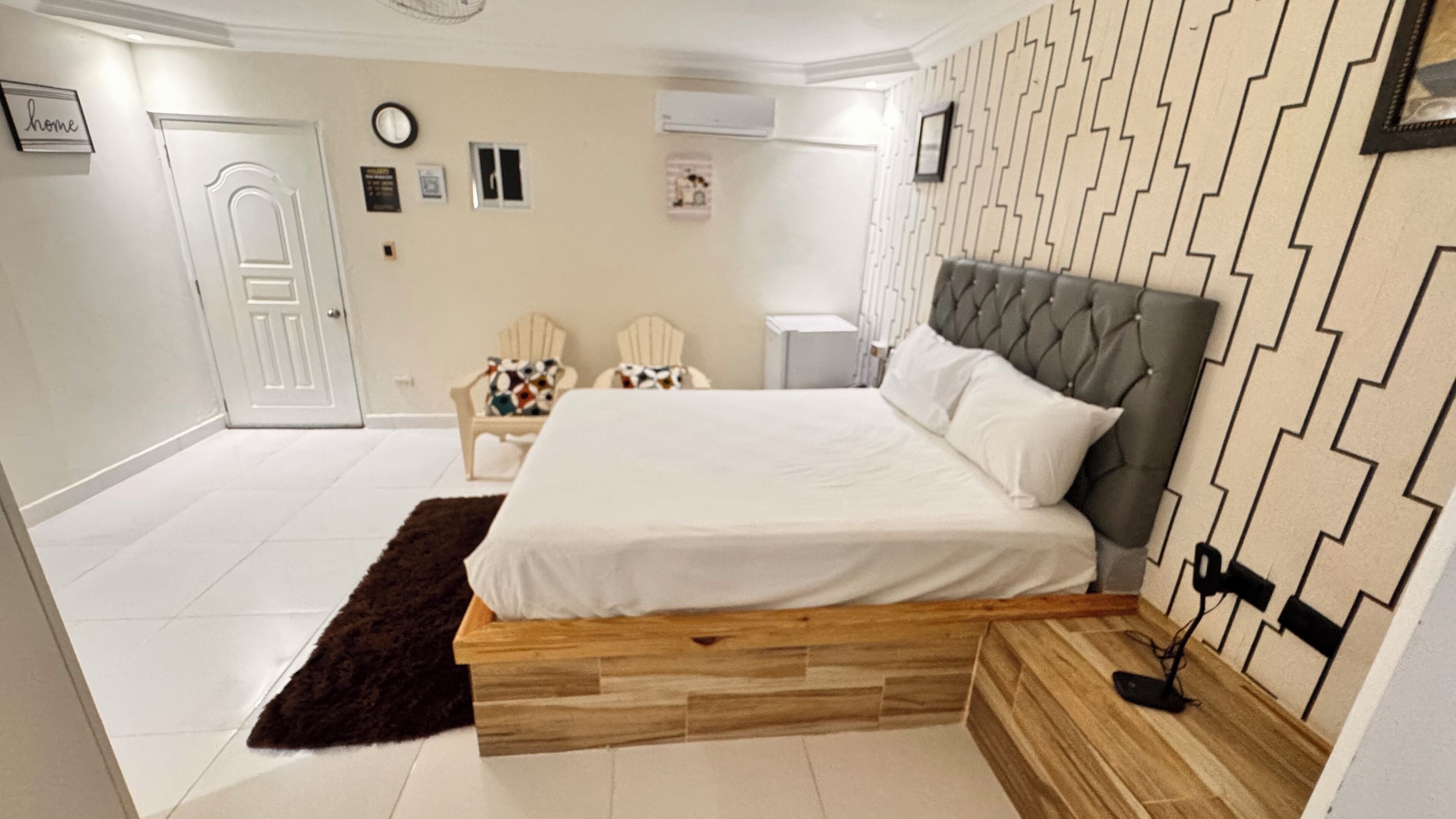 Une chambre à coucher comprenant un lit, une chaise, une table de chevet, une horloge et un climatiseur fixé au mur.
