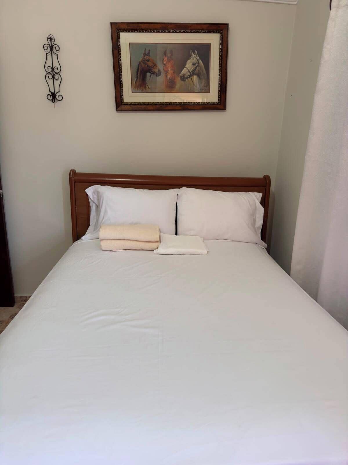 Ein ordentlich gemachtes Bett mit weißen Leinentüchern und zwei Kissen, ein gerahmtes Bild von Pferden an der Wand und ein dekoratives Metallobjekt auf der linken Seite.
