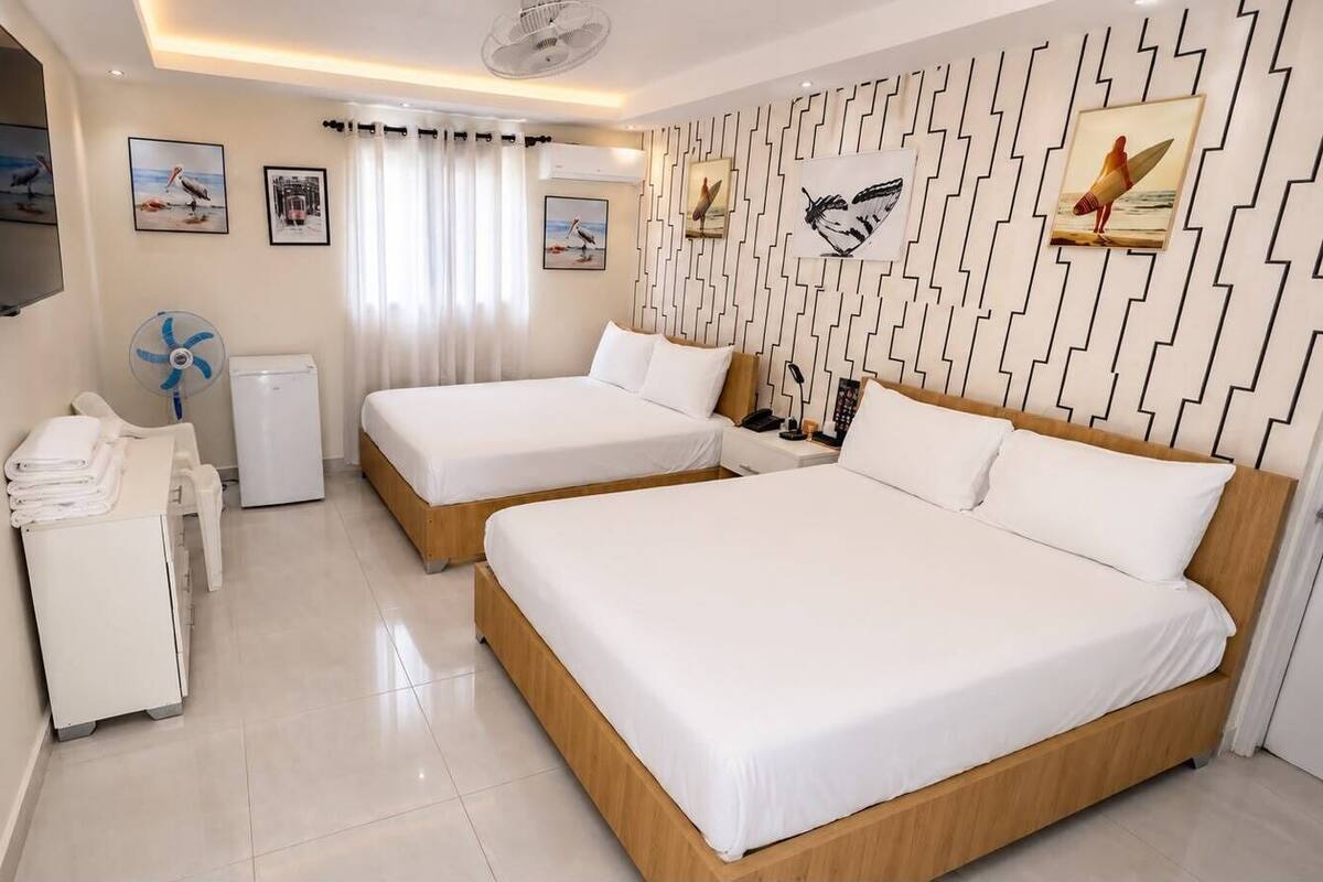 A hotel room with two beds, a flat-screen TV, a desk with a chair, a fan, and artwork on the walls.