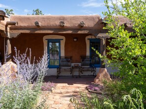Un bâtiment rustique doté d’un patio en pierre, de deux fenêtres à cadre bleu et d’une table en bois avec des chaises.