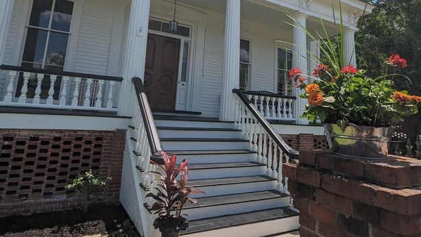 A white colonial-style building with a front porch, brick steps, and a small garden area with potted plants.