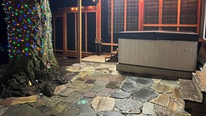A hot tub under a wooden pergola with string lights, surrounded by stone tiles and a tree with colorful lights. - Private Lake House (Puyallup)