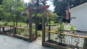 A wooden pergola with flowers, a gravel path, and a fenced garden area with a small American flag. - Private Lake House (Puyallup)