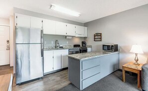 Una cocina compacta con armarios blancos, un refrigerador de acero inoxidable, un microondas, una cafetera y una pequeña mesita de madera con una lámpara.