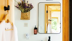 A modern bathroom with a wooden vanity, a white sink, a mirror, and a basket of flowers. - The Cabins at Reverence Farms by Sagra (Graham)