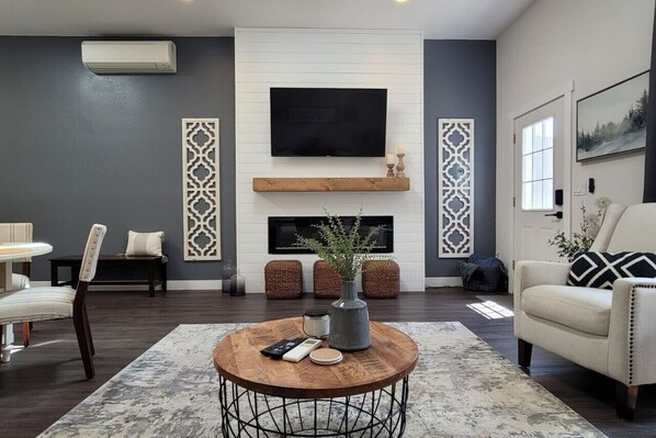 A modern living room with a flat-screen TV mounted above a fireplace, a round coffee table with decorative items, and a comfortable seating area with a sofa and chairs.