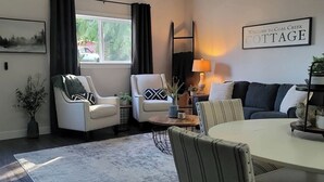 A cozy living room with a sofa, two armchairs, a dining table, and a coffee table. There is a window with curtains, a lamp, and a sign that reads "Welcome to Coal Creek Cottage." - Coal Creek Cottage (hot tub, dog and kid friendly) (Sedro-Woolley)