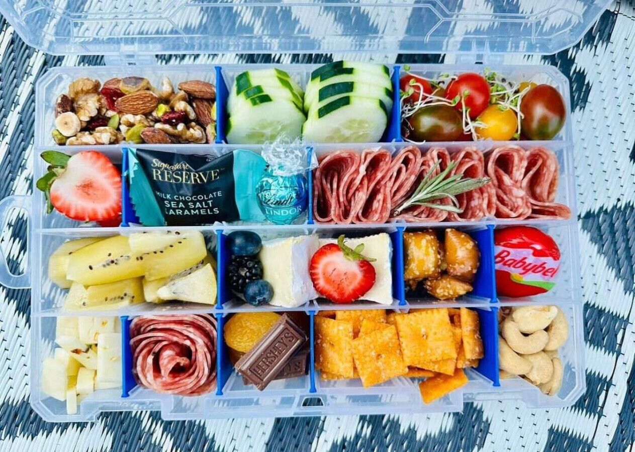 Un recipiente para alimentos con varios compartimentos que contienen nueces, pepinos en rodajas, tomates cherry, fresas, queso y galletas.