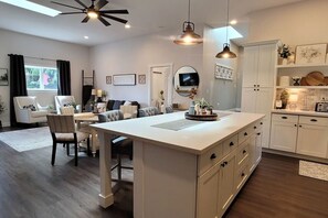 A modern kitchen with white cabinetry, a central island, pendant lighting, and a dining area with a sofa and chairs.