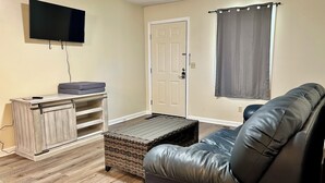 Una sala de estar con un sofá de cuero, un televisor de pantalla plana montado en la pared, un mueble de madera con estantes y un reposapiés de mimbre.