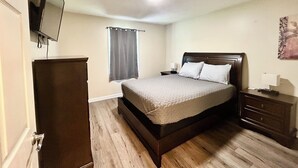 Un dormitorio con cama, cómoda, mesita de noche, televisor montado en la pared y ventana con cortinas.