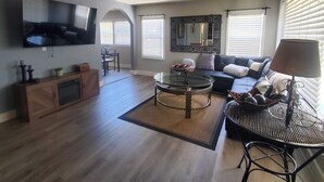 A spacious living room with a flat-screen TV mounted on the wall, a dark-colored sofa, a round coffee table, and a dining area with a wooden cabinet and chairs. - 2/1 Cozy Home in the Heart of the City (Stockton)