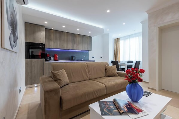 A modern living room with a beige sofa, a coffee table with magazines, and a blue vase with red flowers.
