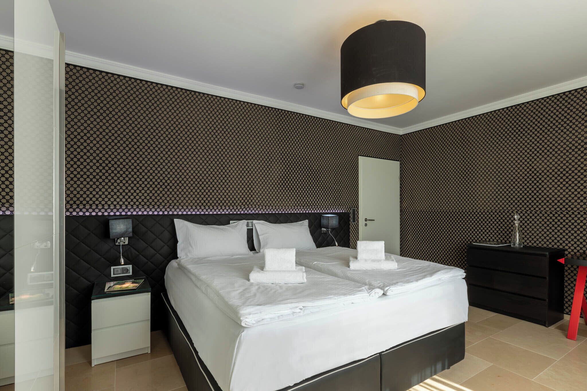 Een moderne hotelkamer met een zwart-wit behang, een groot bed met witte lakens, een nachtkastje met een lamp en een rode kruk.