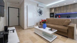 Un salon moderne avec un canapé beige, une table basse blanche ornée d’un vase de fleurs rouges, et un coin cuisine doté de meubles en bois et d’un éclairage LED bleu.