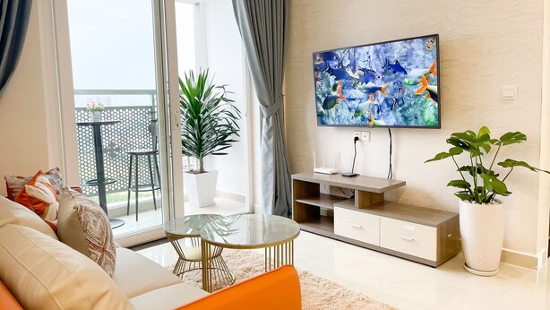 Una sala de estar moderna con un sofá, una mesa de centro de vidrio, un televisor montado en la pared y un balcón con barandilla y plantas en macetas.