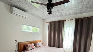 Une chambre à coucher avec un lit, une table de chevet, un ventilateur de plafond, une fenêtre et des rideaux.