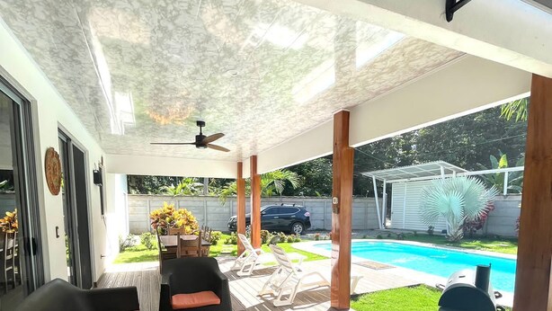 Une terrasse couverte, aménagée avec des meubles en osier, dotée d’un ventilateur de plafond et offrant une vue sur une piscine et un jardin.