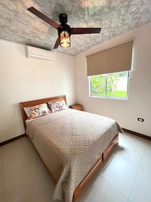 Une chambre avec un lit, un ventilateur de plafond, une fenêtre donnant sur un espace verdoyant et un climatiseur fixé au mur.