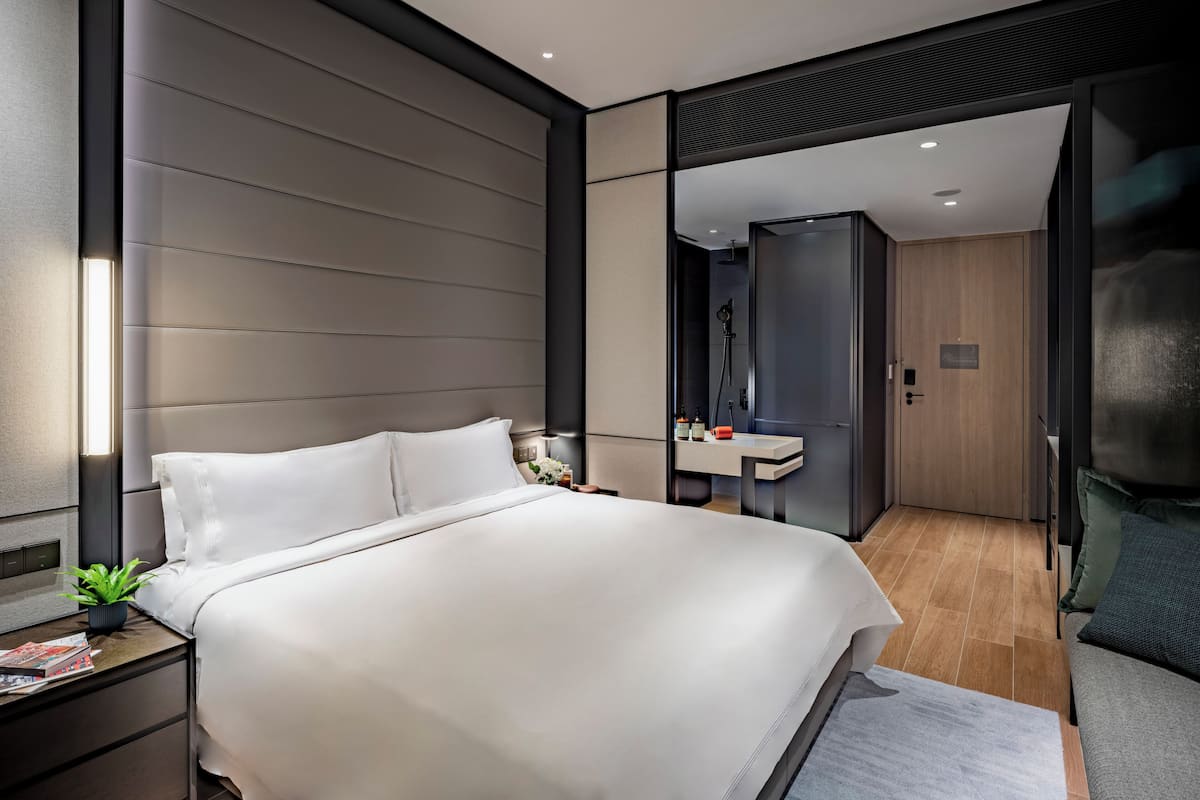 A modern hotel room with a large bed, bedside table, and a small plant. There is a bathroom visible through a glass door.