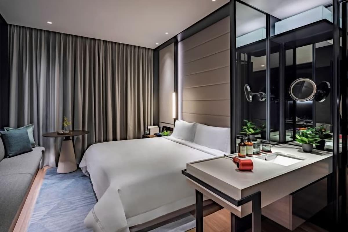 A modern hotel room with a large bed, bedside tables, a desk with a mirror, and a view of the city through floor-to-ceiling windows.