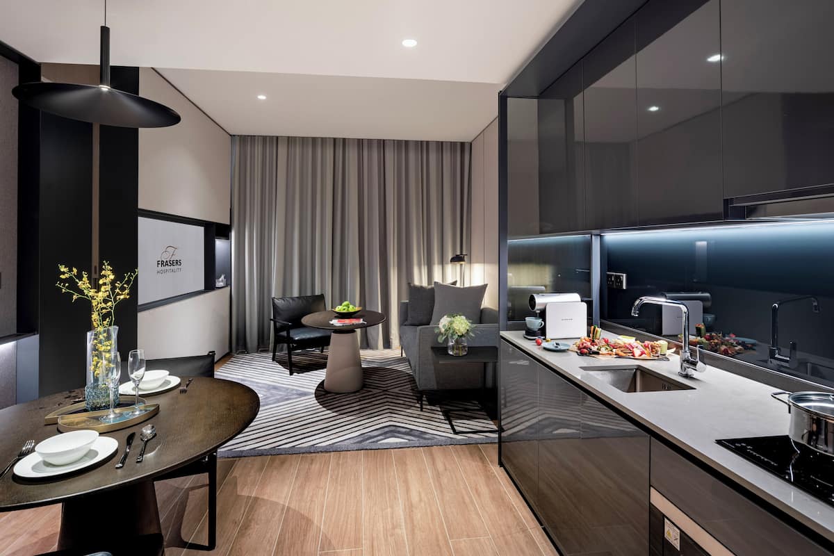 A modern hotel room with a dining area, a sofa, a kitchen with a built-in oven, and a large window with curtains.