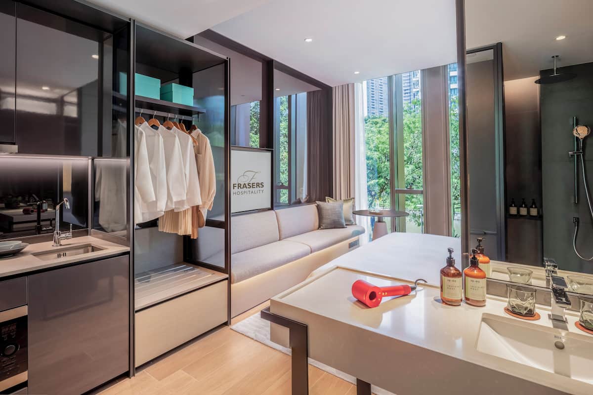 A modern hotel room with a kitchenette, a seating area with a sofa, a large window with a city view, and a bathroom with a shower and toiletries.