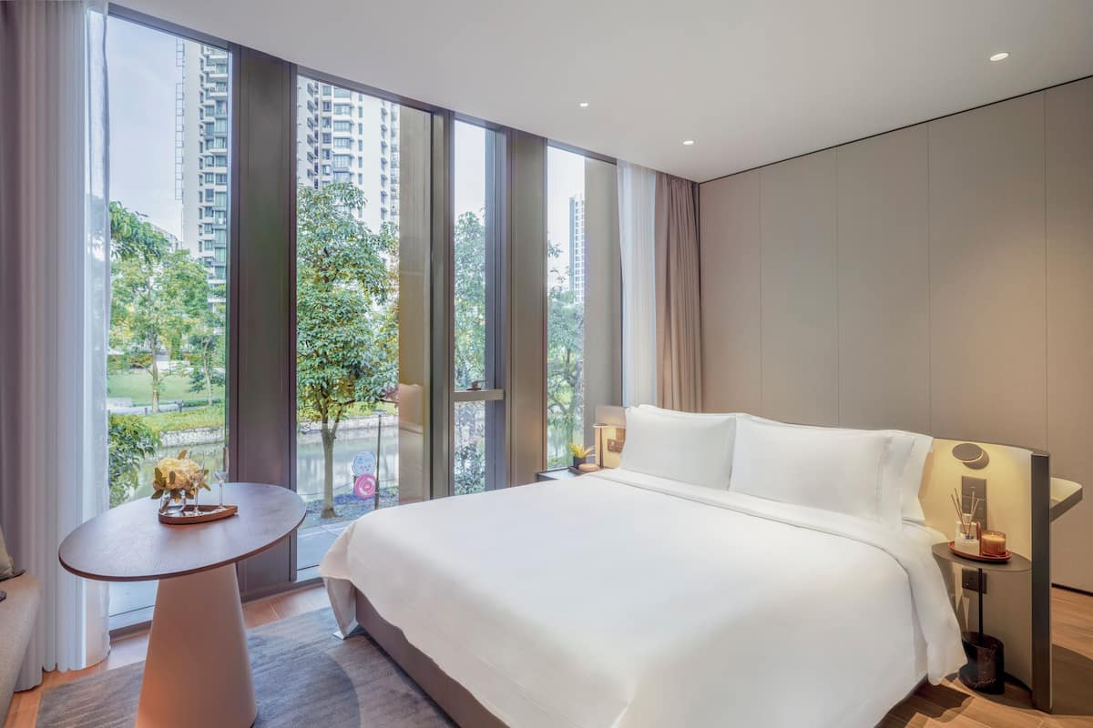 A modern hotel room with a large bed, a round table, and a view of a cityscape through large windows.