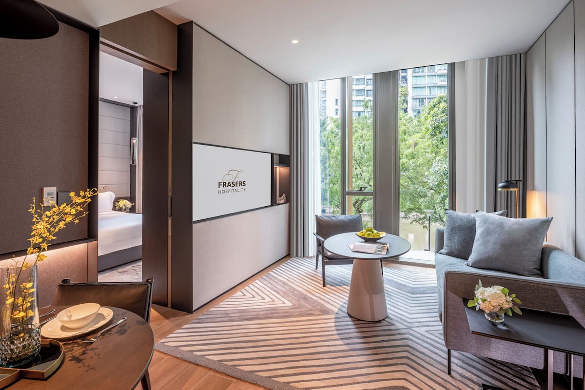 A modern hotel room with a large window, a sofa, a dining table, and a small balcony.