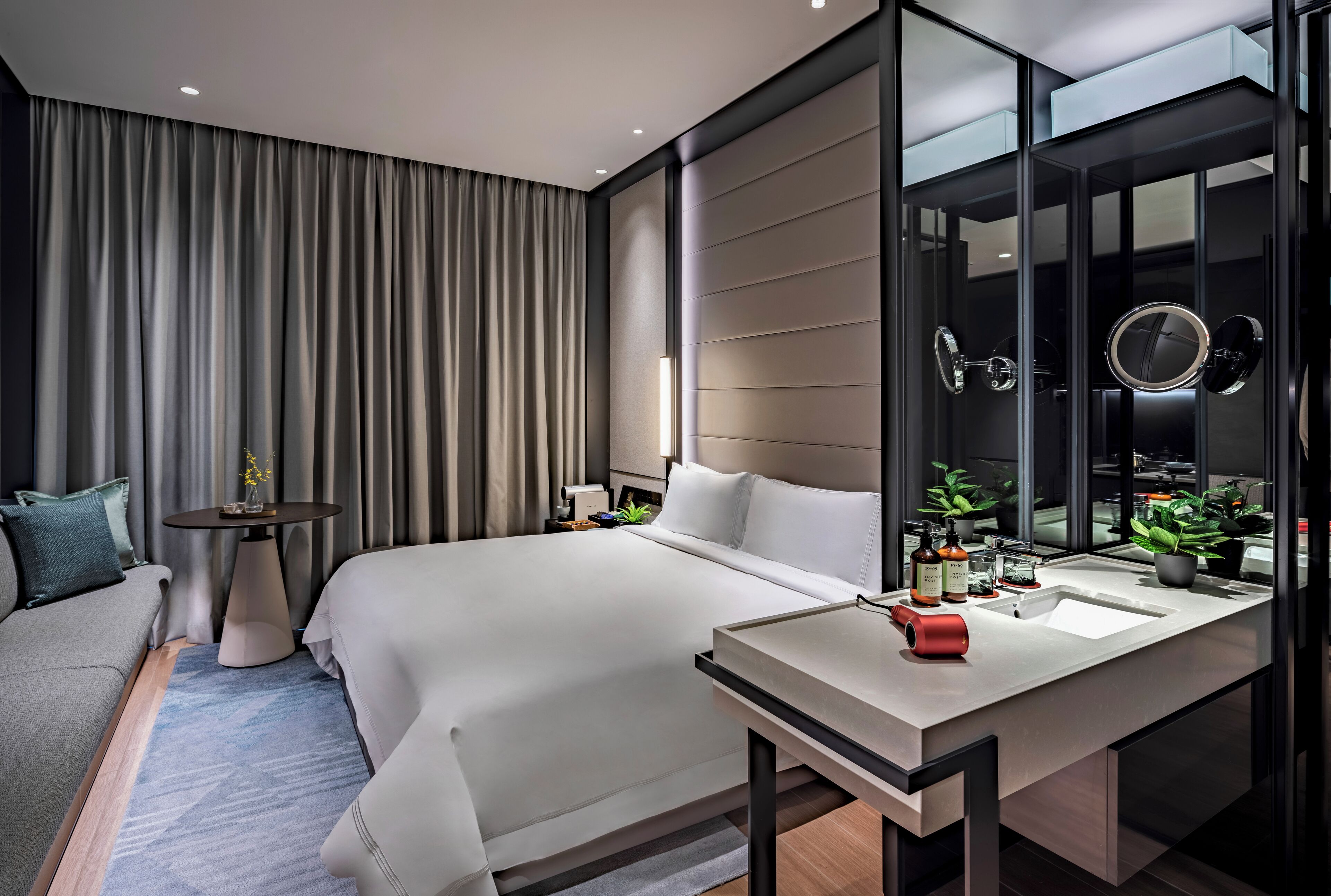 A modern hotel room with a large bed, a desk with a sink, a mirror, and a view of the city through large windows.