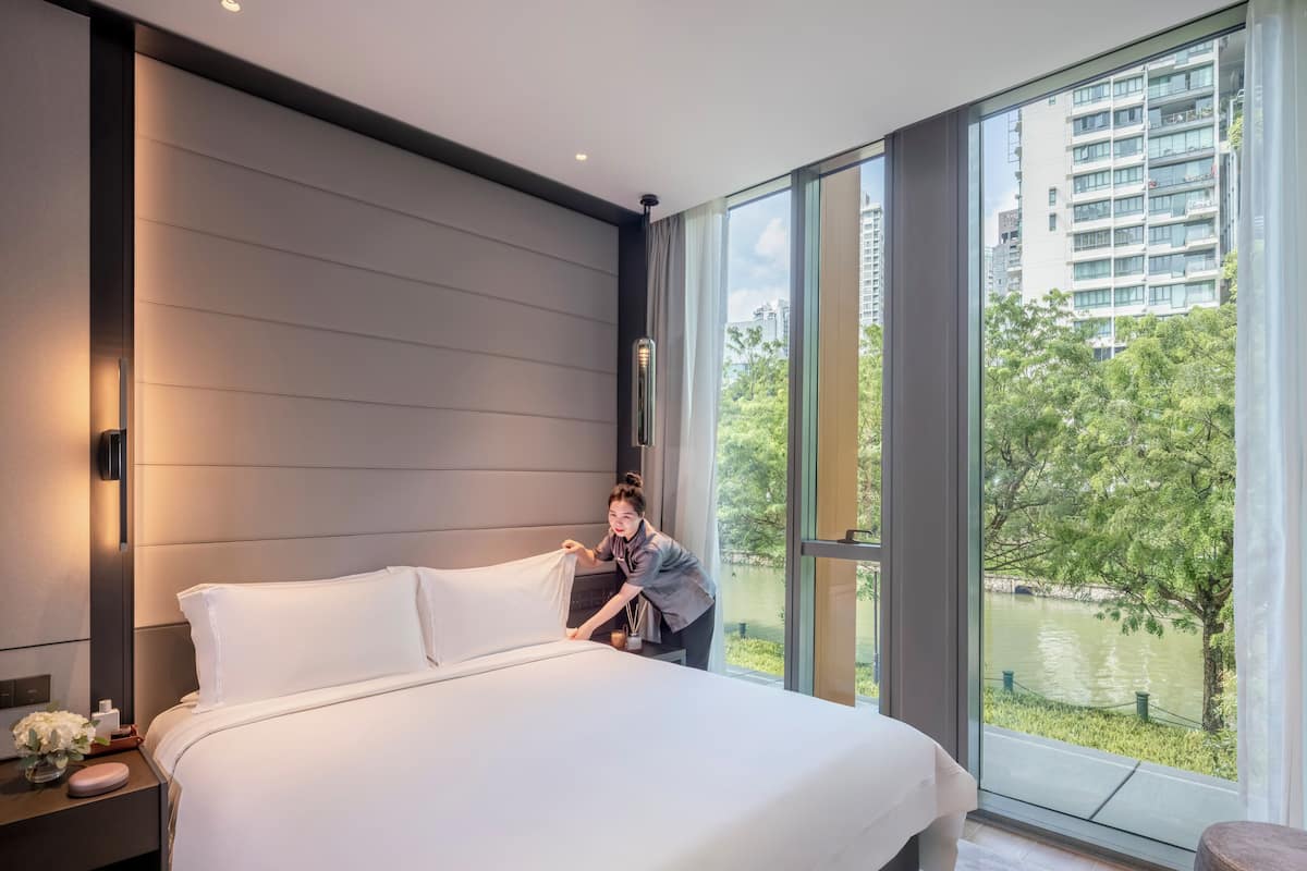 A modern hotel room with a large bed, a nightstand with a vase of flowers, and a view of a river and buildings through large windows.