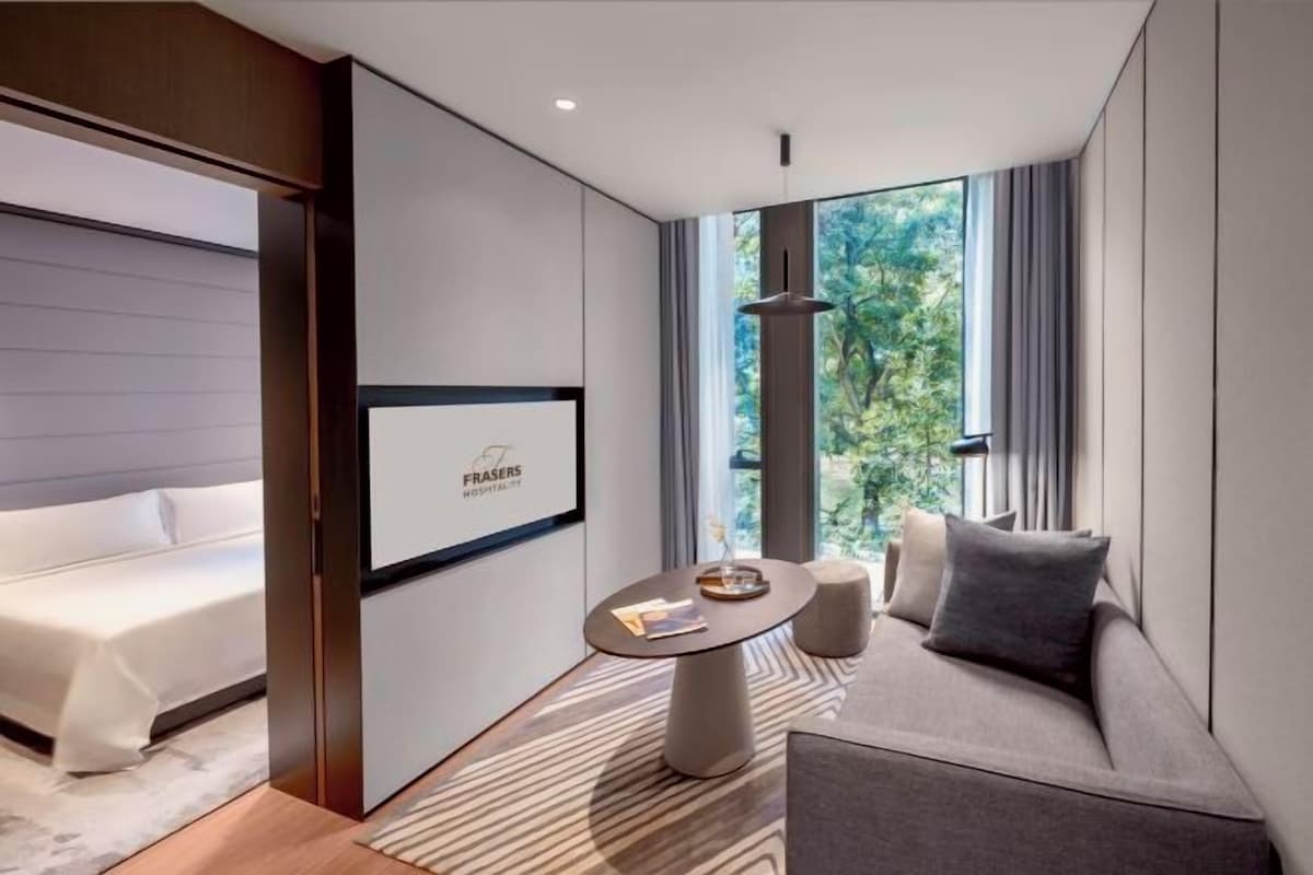 A modern hotel room with a bed, a sofa, a small table, and a large window offering a view of greenery.
