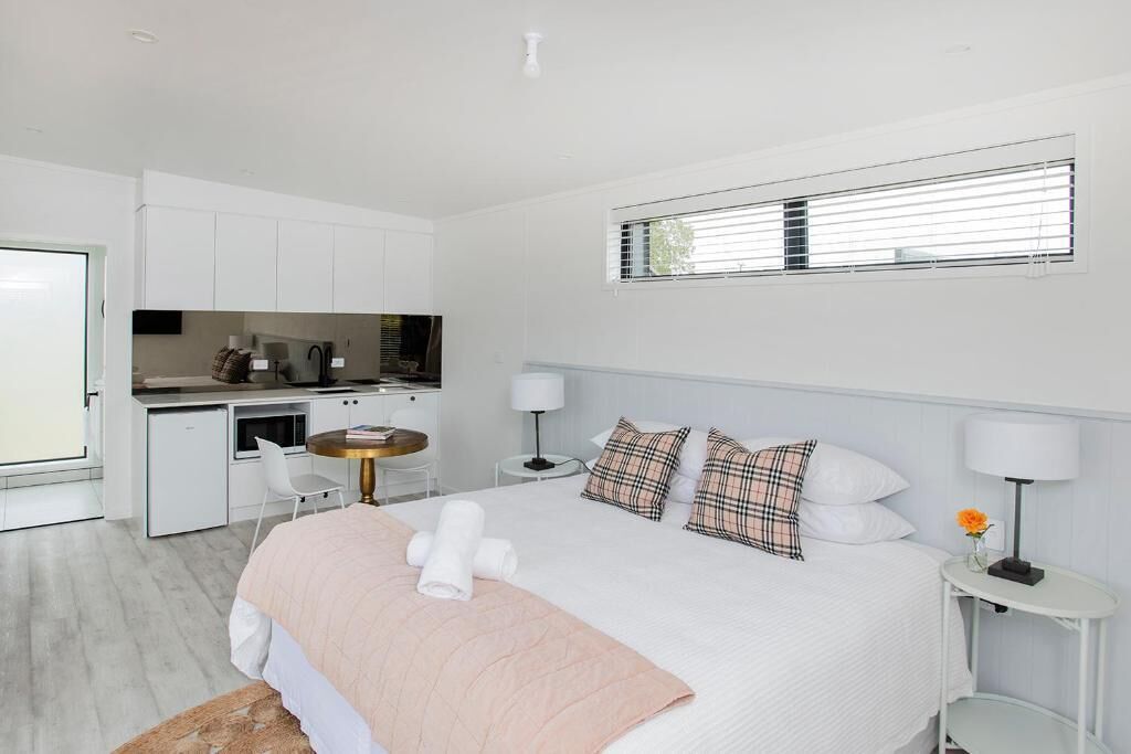A modern hotel room with a large bed, a small kitchenette, a dining area, and a window with blinds.