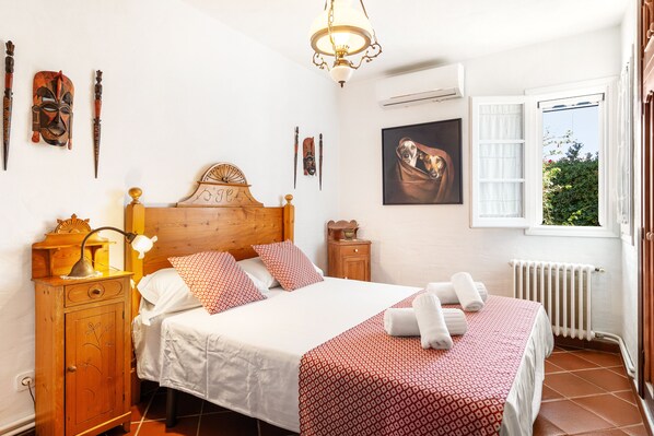 Un dormitorio ordenado con cabecera de madera, cama con ropa de cama blanca y roja, mesita de noche con lámpara y ventana con persiana abierta.