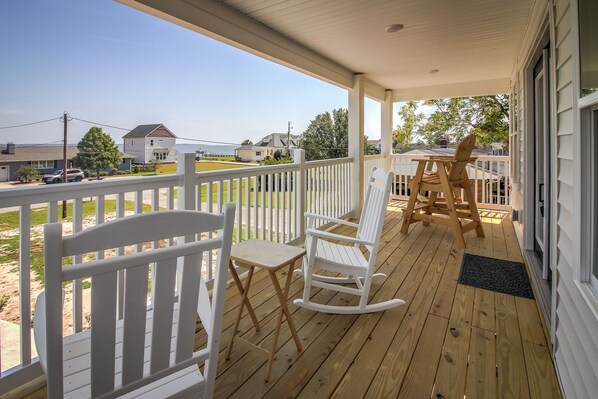 A wooden deck with white rocking chairs and a table, overlooking a grassy area and houses.
