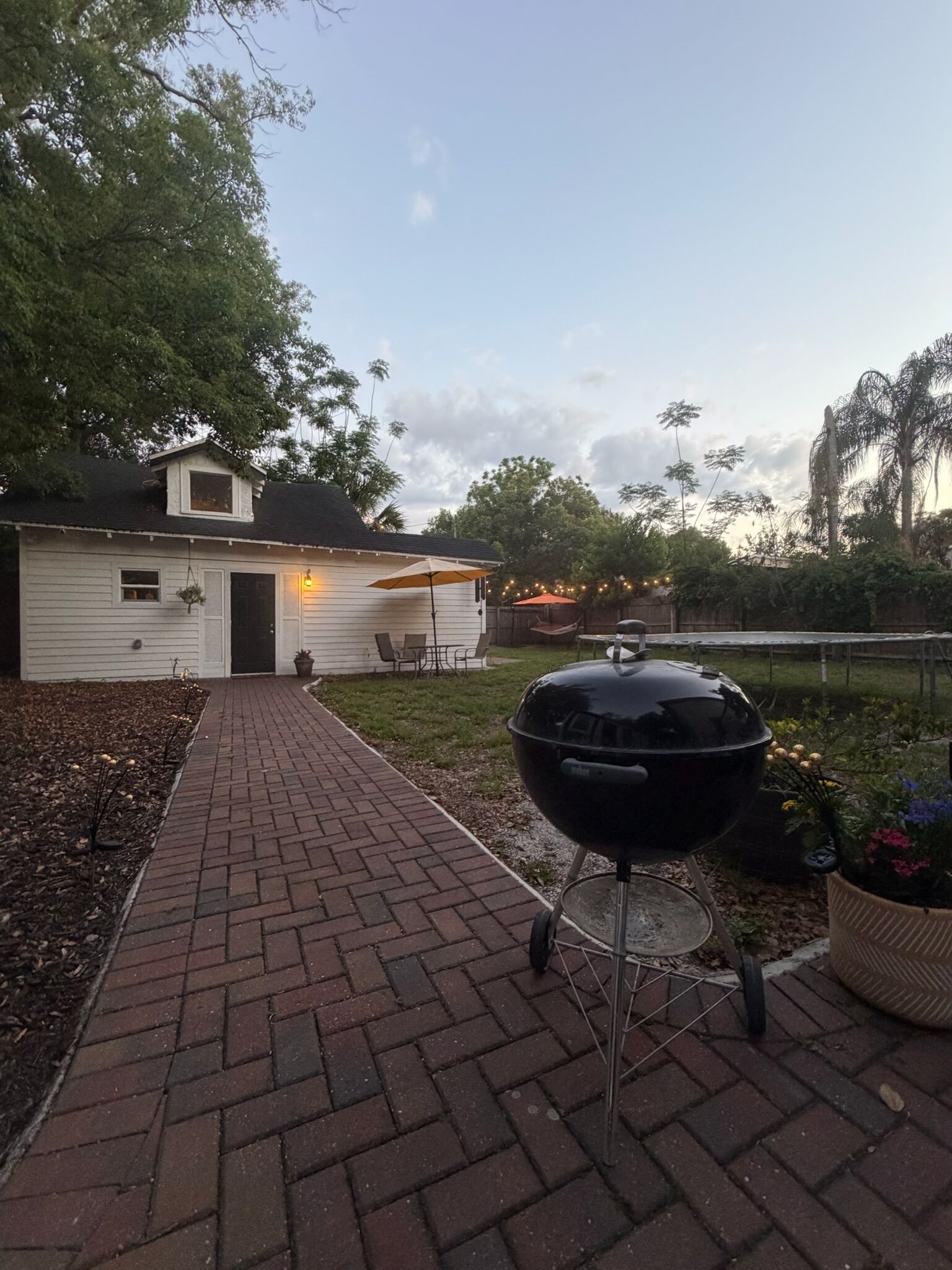 A backyard with a brick pathway leading to a white house, a black grill, and outdoor furniture.