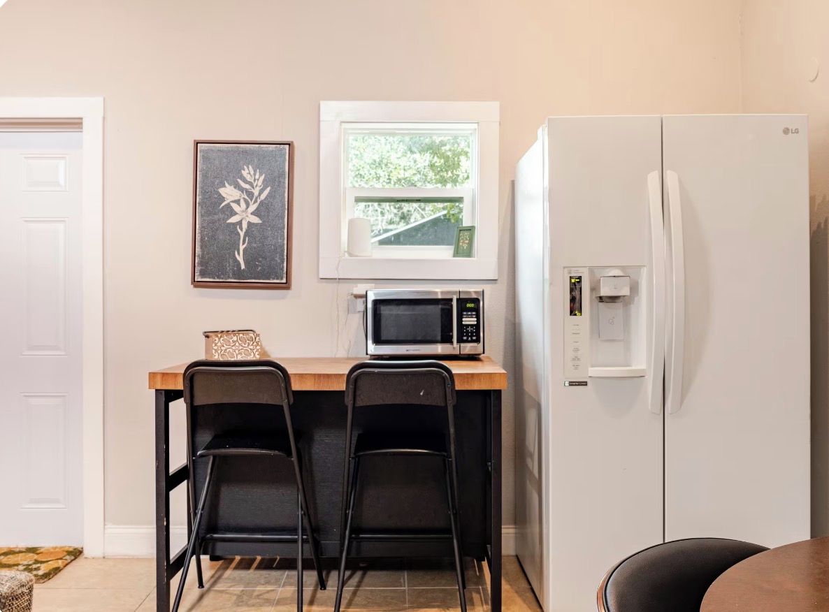A compact kitchen with a microwave, two chairs, a refrigerator, a window, and a door.