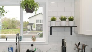 A kitchen with white tiles, a hanging plant, a window with a view of a house, and a shelf with potted plants. - Renovated Hidden-Gem Minutes from Downtown Denver with coffee bar (Aurora)