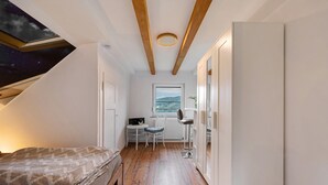 Une petite pièce comprenant un lit, un bureau, un radiateur et une fenêtre donnant sur des collines.