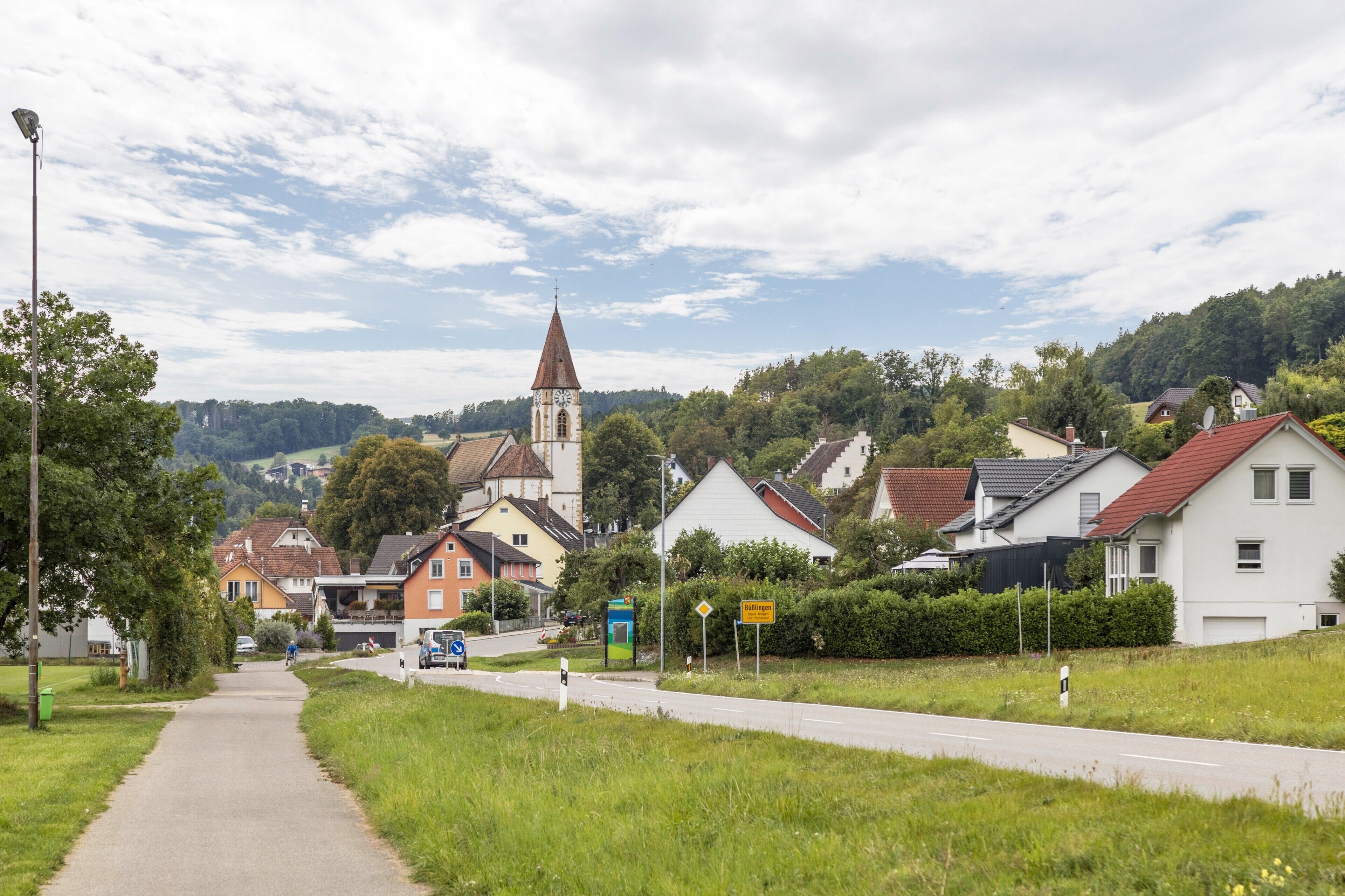 A rural road leading to a village with houses, a church, and a sign indicating a nearby town.