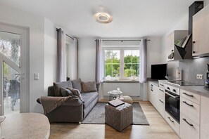 A modern kitchen with a built-in oven, a dining area with a round table, and a sofa with a view of greenery outside.